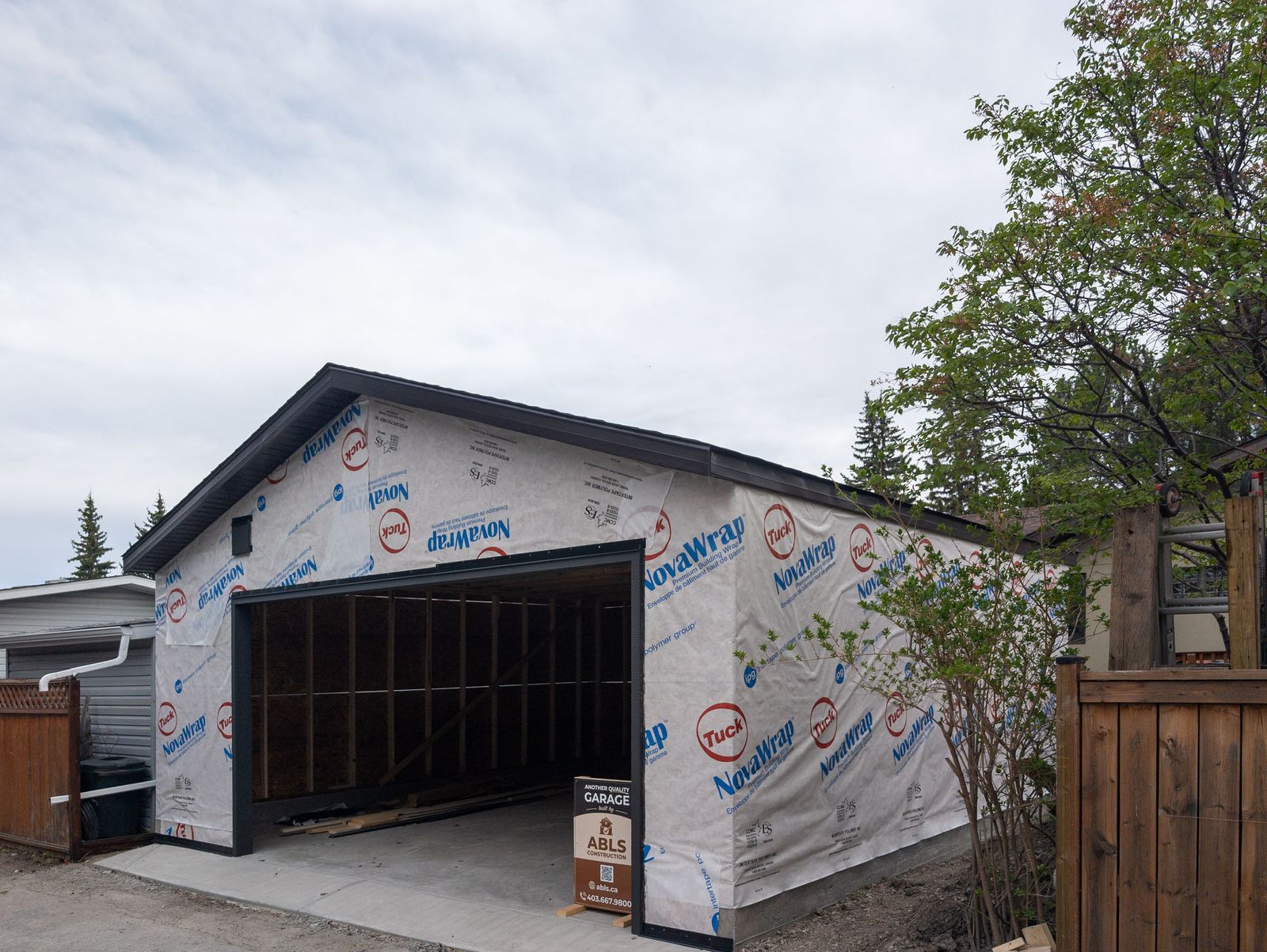 The garage framed and wrapped in house wrap, with the ABLS sign out front and the poured pad and apron