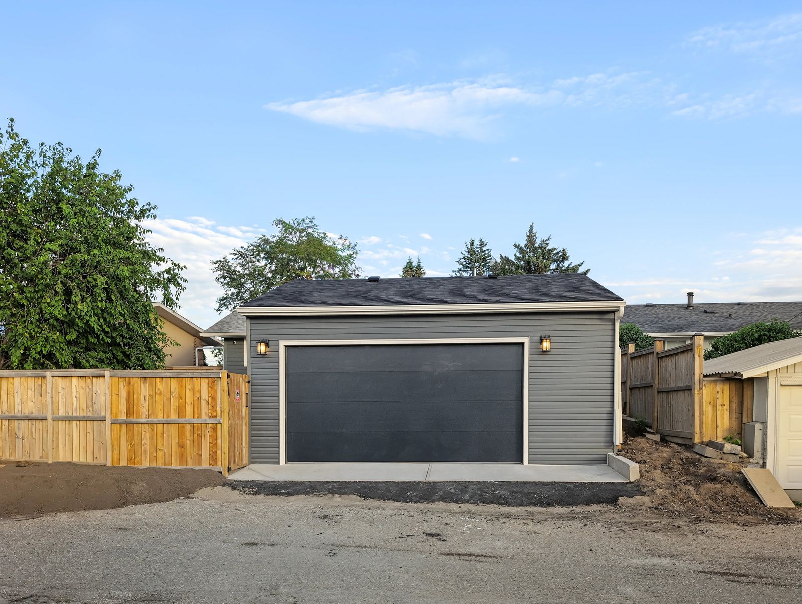 The finished garage from across the lane: dark grey siding, black overhead door, new cedar fence on the left