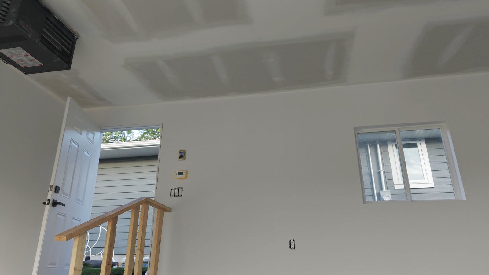 Inside the garage: taped drywall walls and ceiling, the gas unit heater hung in the corner, the thermostat, and steps with a railing up to the open man door