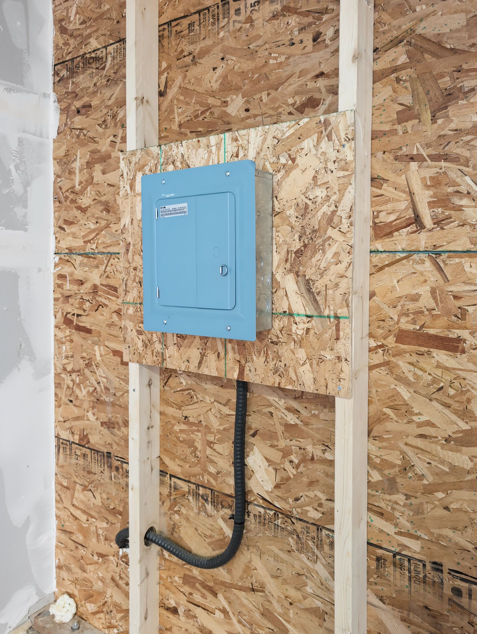 The garage sub panel, a blue-covered load centre mounted on a plywood backer between studs, with flexible conduit running down into the wall
