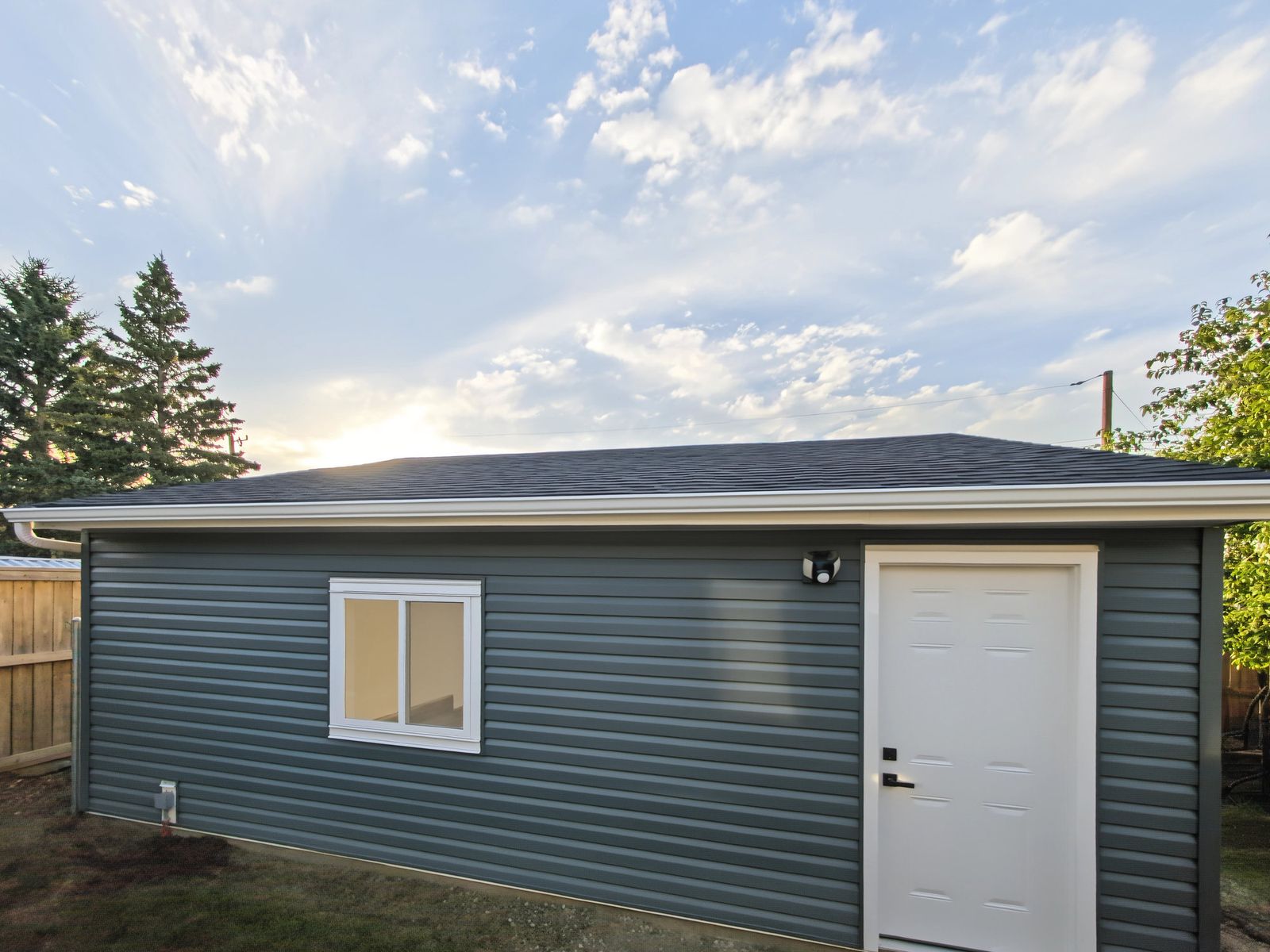 The garage from the back yard: sliding window, white man door and a motion light under the eave, with the exterior receptacle low on the wall