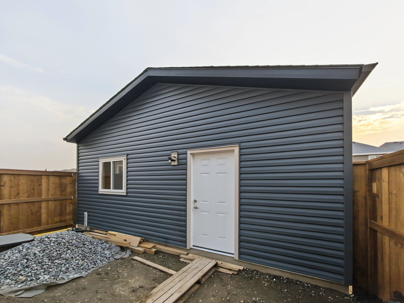 The garage from the corner of the yard, showing the side wall, the man door and the gravel base along the wall