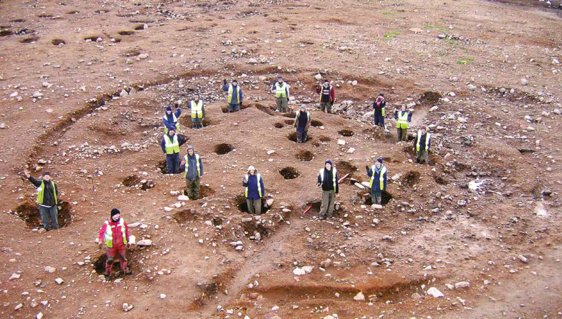 People on an archaeological site standing inside postholes which would have formed a roundhouse