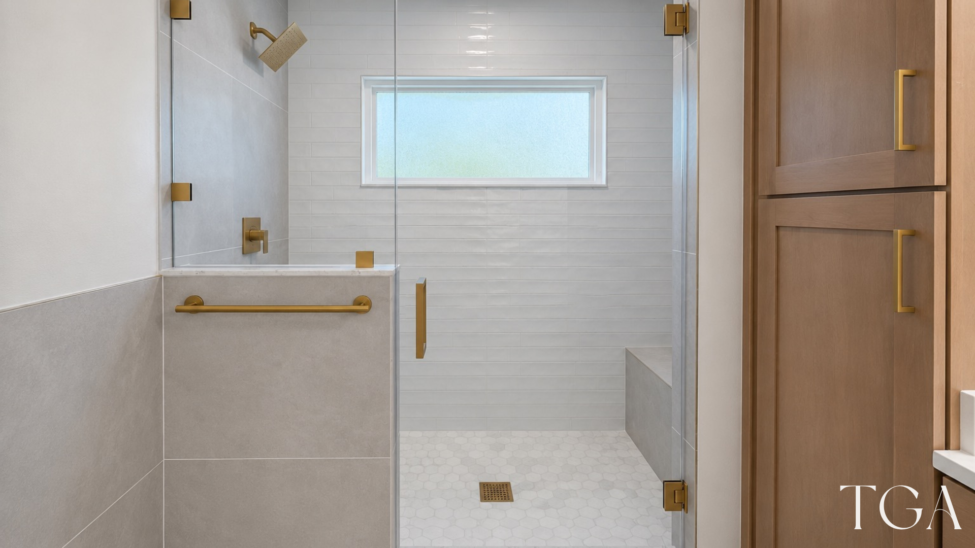Glass shower enclosure with brushed gold hardware, white wall tile, a built-in bench and hexagon floor tile.