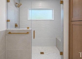 Glass shower enclosure with brushed gold hardware, white wall tile, a built-in bench and hexagon floor tile.
