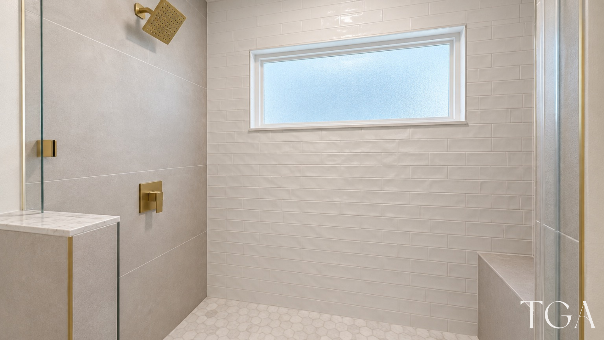 Brushed gold square showerhead above white hexagon floor tile, with gray side tile and a shower bench.
