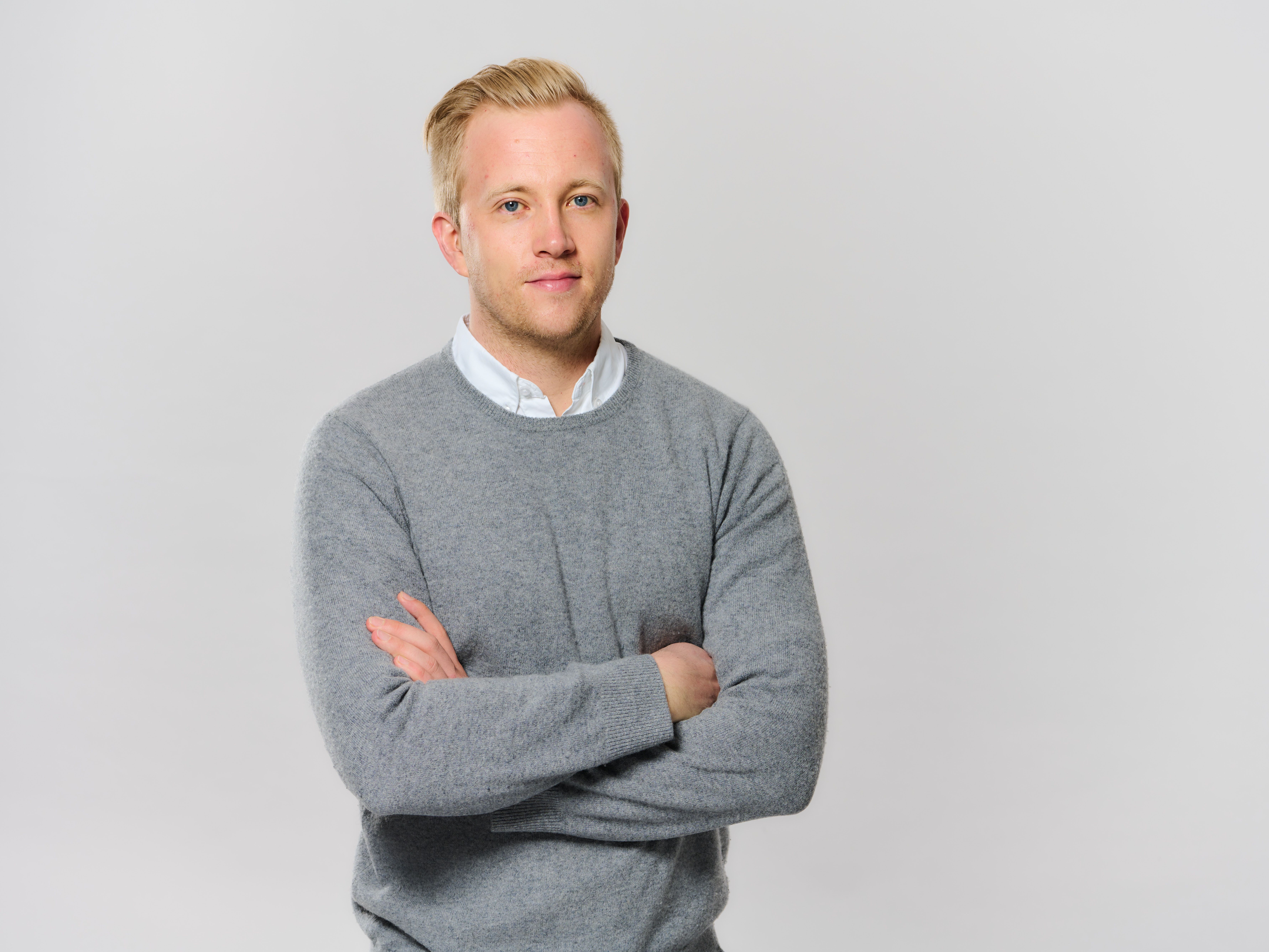 Person mit blondem Haar steht vor hellem Hintergrund, trägt einen grauen Pullover über einem weißen Hemd und blickt mit verschränkten Armen in die Kamera.