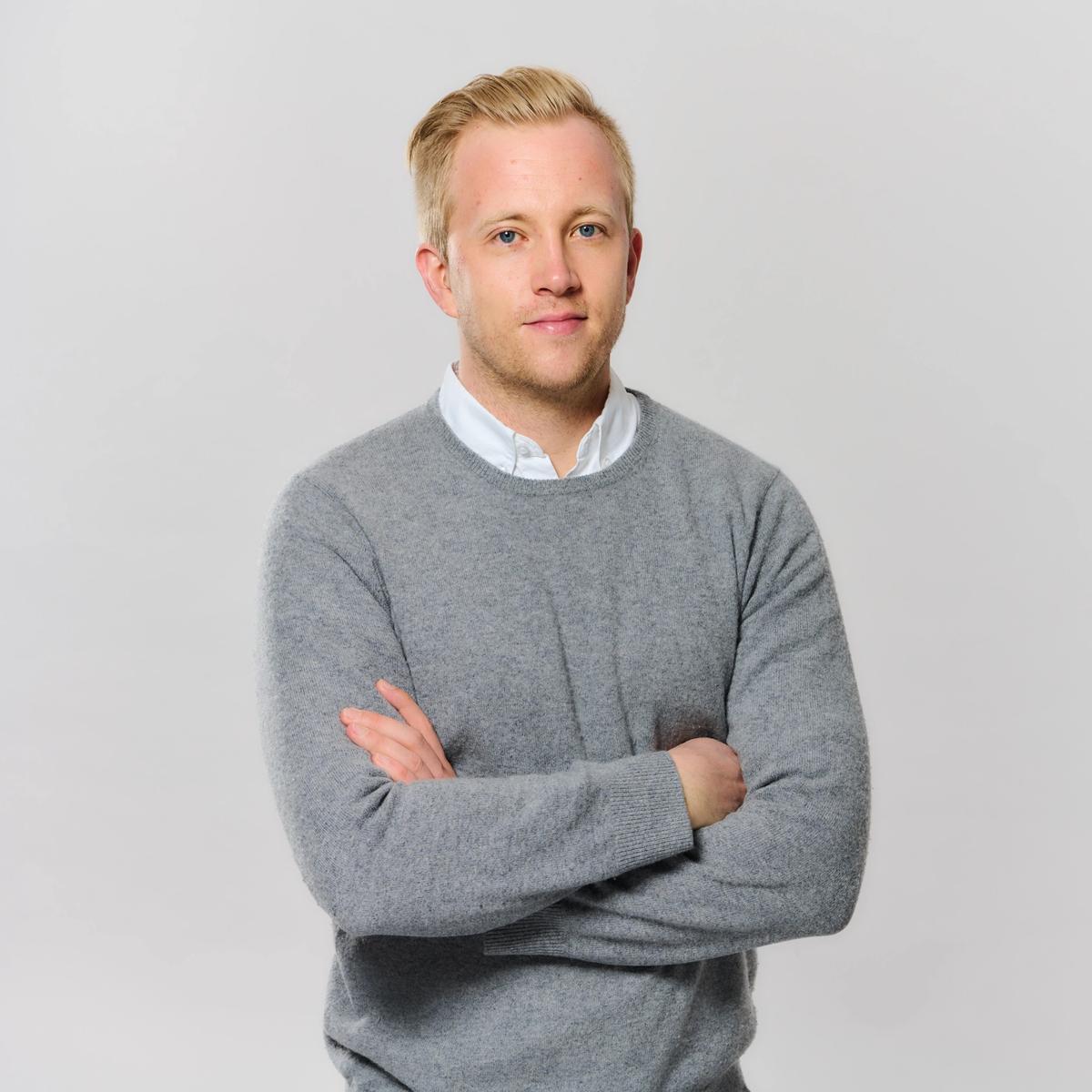 Person mit blondem Haar steht vor hellem Hintergrund, trägt einen grauen Pullover über einem weißen Hemd und blickt mit verschränkten Armen in die Kamera.