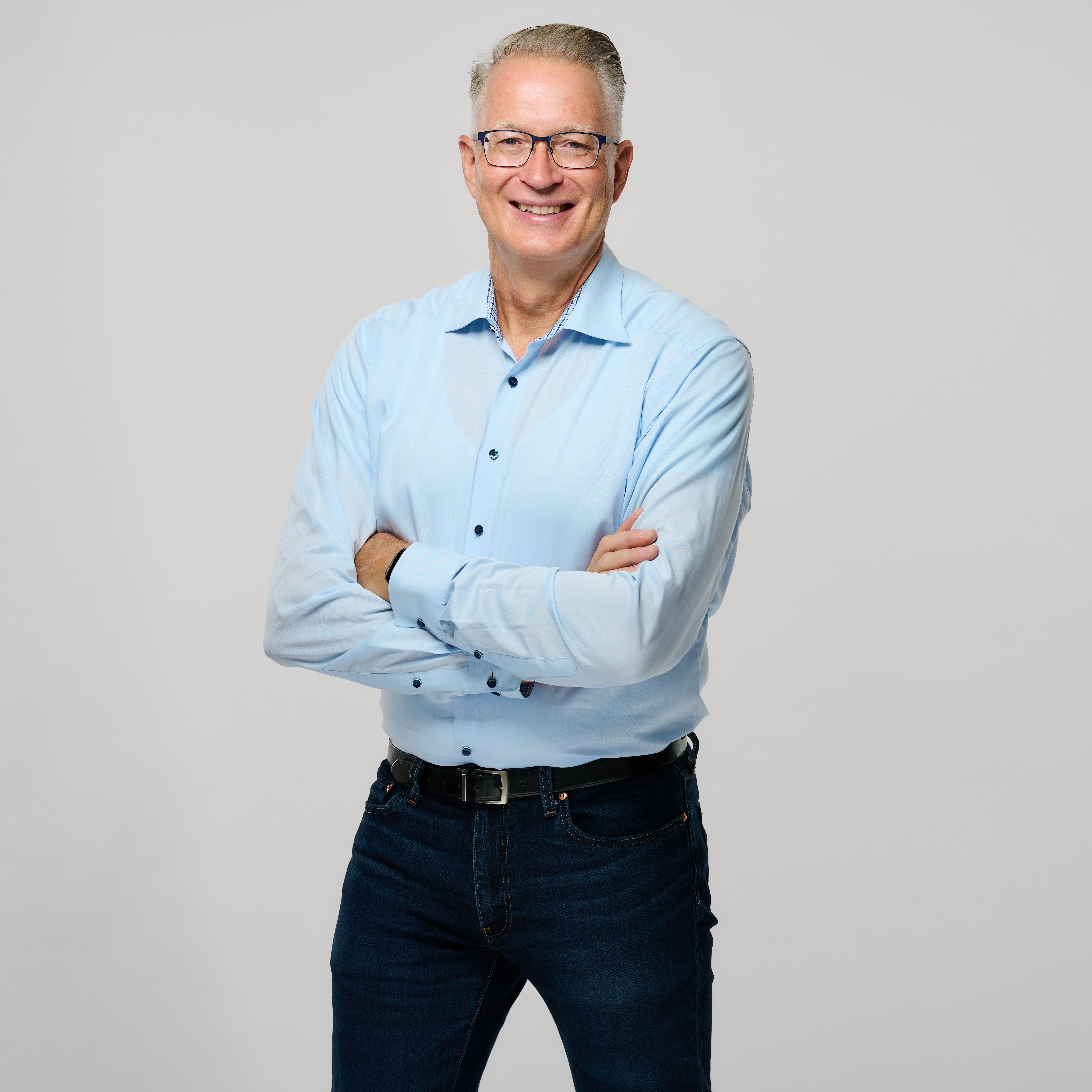 Person mit kurzem, grauem Haar und Brille steht vor einem hellen Hintergrund, trägt ein hellblaues Hemd und dunkle Jeans und lächelt mit verschränkten Armen in die Kamera.