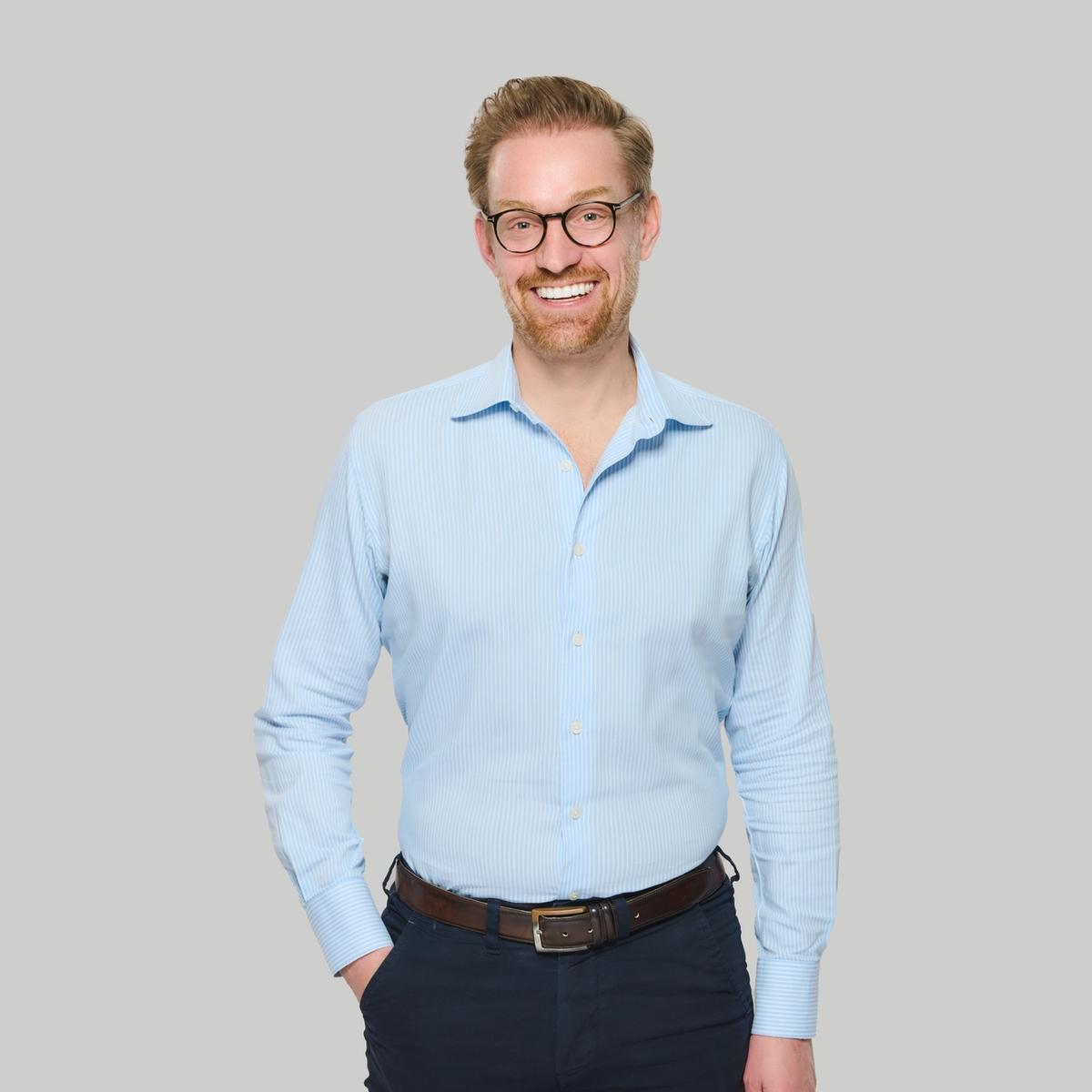 Person mit kurzem blondem Haar und Brille steht vor hellem Hintergrund, trägt ein hellblaues Hemd und eine dunkle Hose und lächelt in die Kamera.