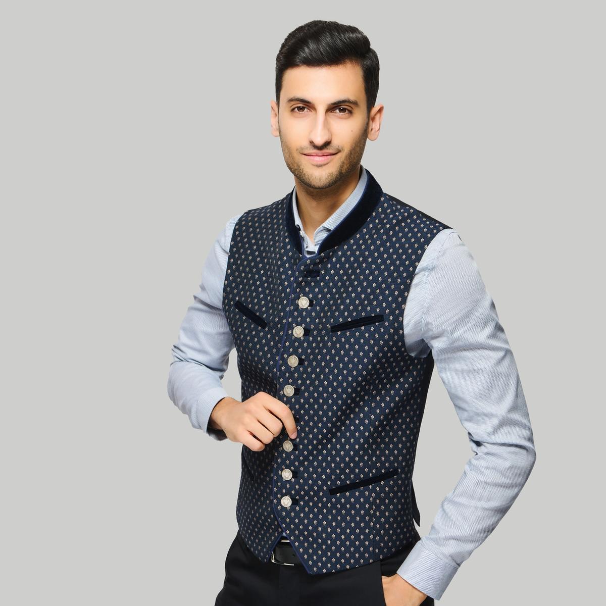 Person mit dunklem Haar steht vor hellem Hintergrund, trägt ein hellblaues Hemd und eine traditionelle dunkelblaue Weste mit Knöpfen und Muster.