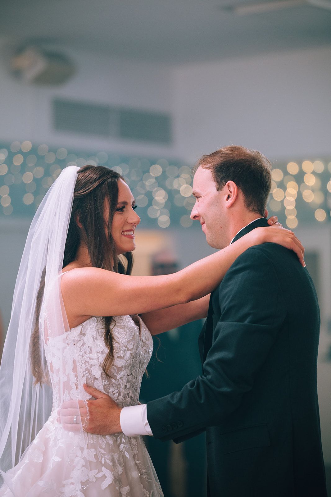 Bride with long dark waves, soft makeup, and a veil smiling at the groom outdoors.