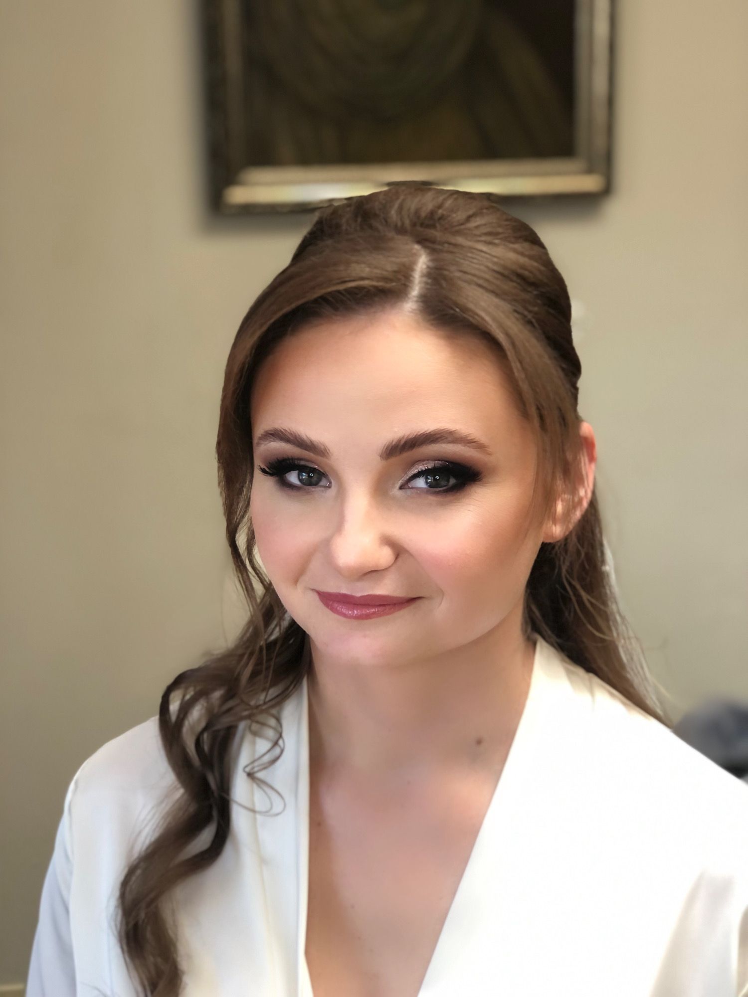 Brunette woman in a white robe with a half-up hairstyle and soft glowing makeup.