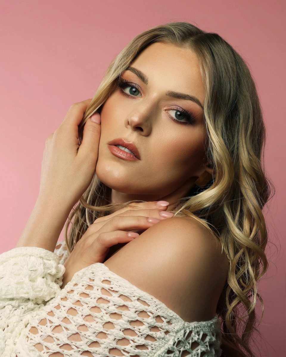 Portret de aproape al unui model blond, cu machiaj al ochilor în tonuri de roz, buze nude și bucle lejere, pe fundal roz.