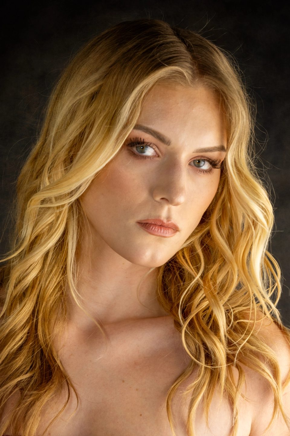 Portrait of a blonde model with loose, face-framing golden waves against a dark background.
