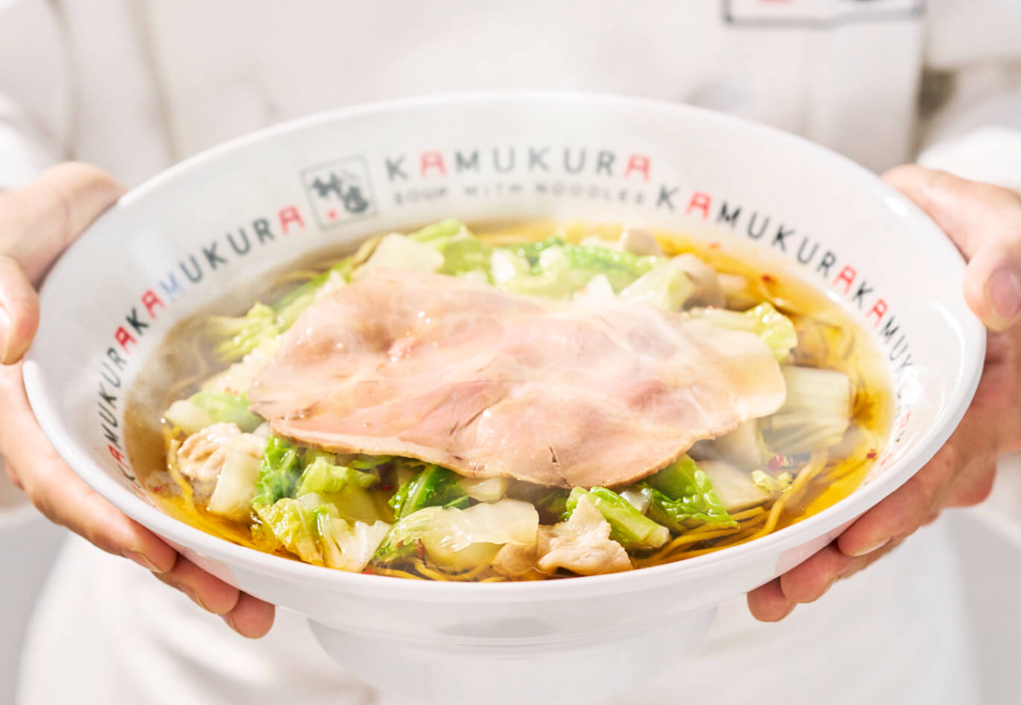 Hands holding a steaming bowl of Kamukura ramen with napa cabbage and a chashu pork slice