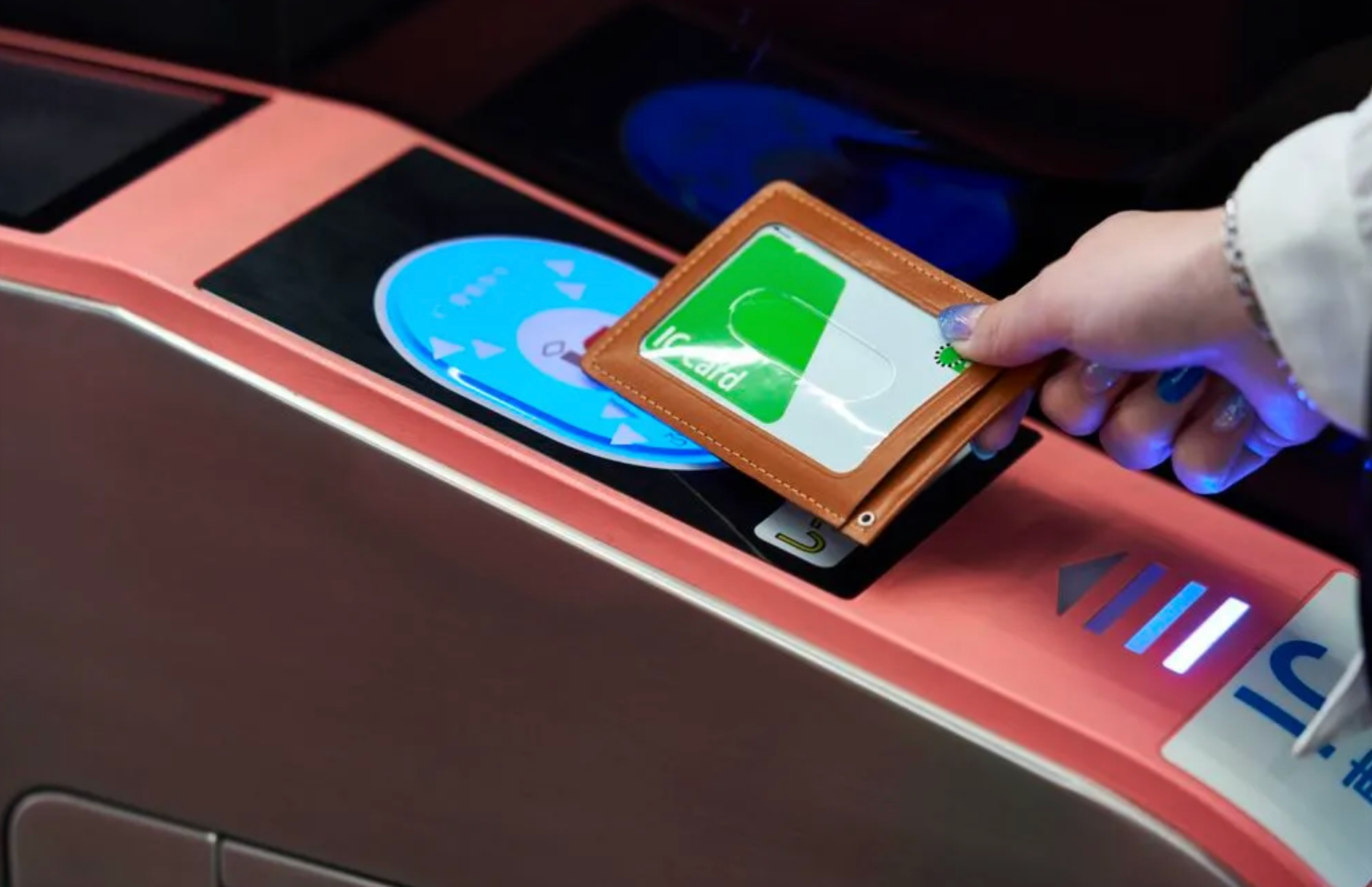 Hand tapping a green IC card in a leather case onto a blue ticket gate reader