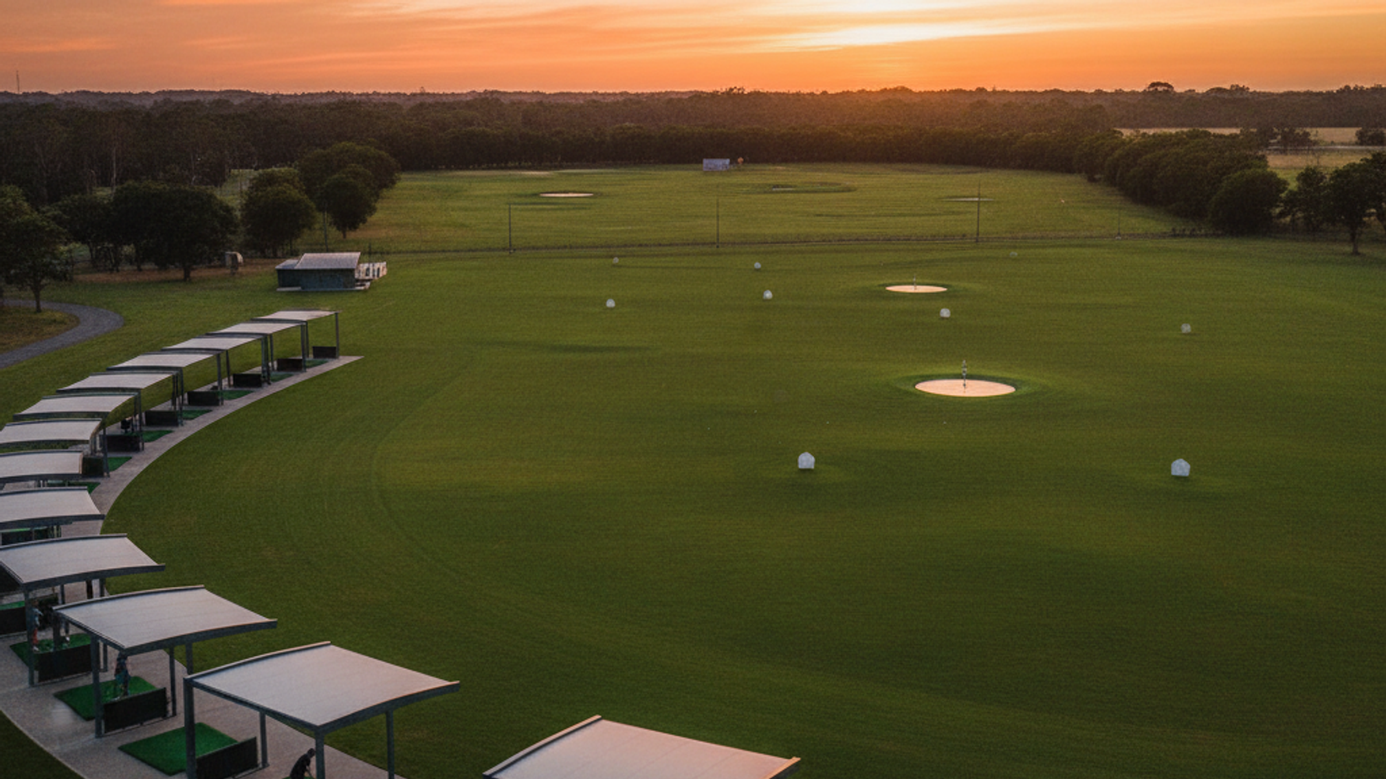 Modern driving range at golden hour with covered hitting bays and golfers practicing