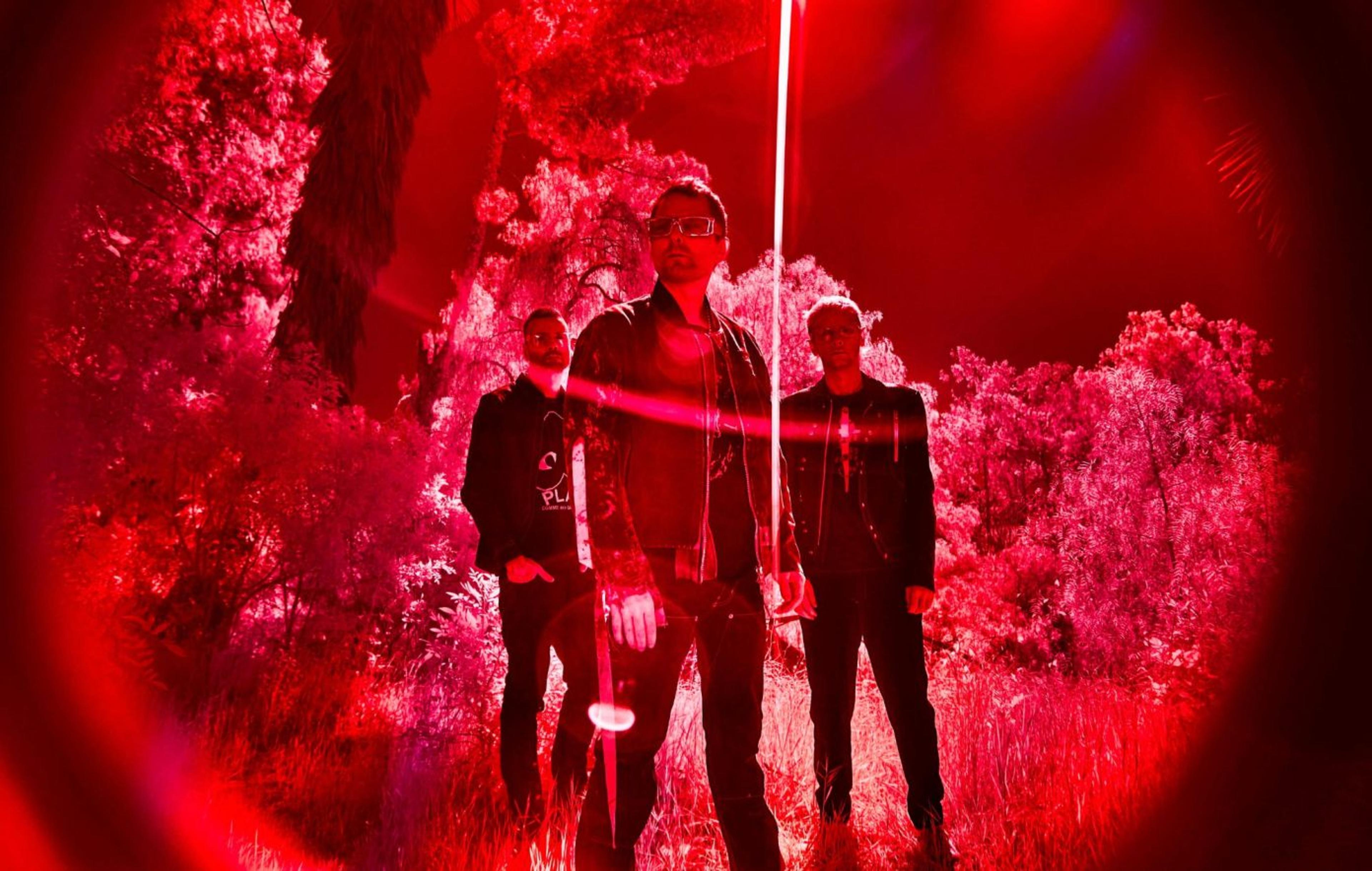 Three band members stand in a vibrant red-lit outdoor setting, surrounded by foliage, exuding a bold and edgy vibe.