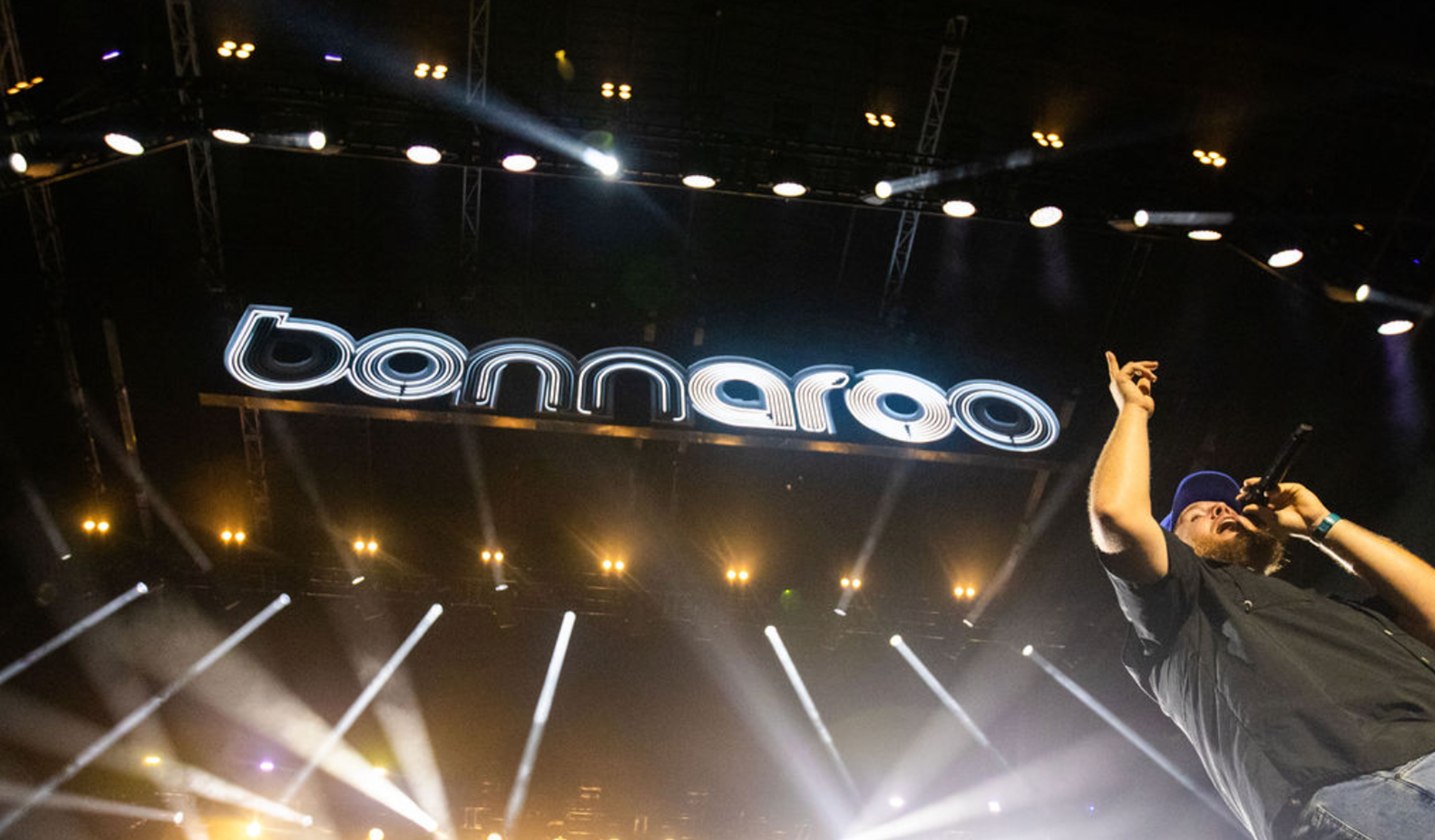 Luke Combs performs on stage at Bonnaroo, with dramatic lighting and the festival's logo prominently displayed above him.