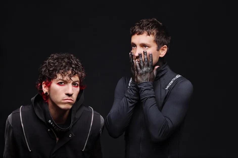 Musicians Twenty One Pilots pose against a black background. One has red-tinted eyes and curly hair, while the other covers his face with hands.