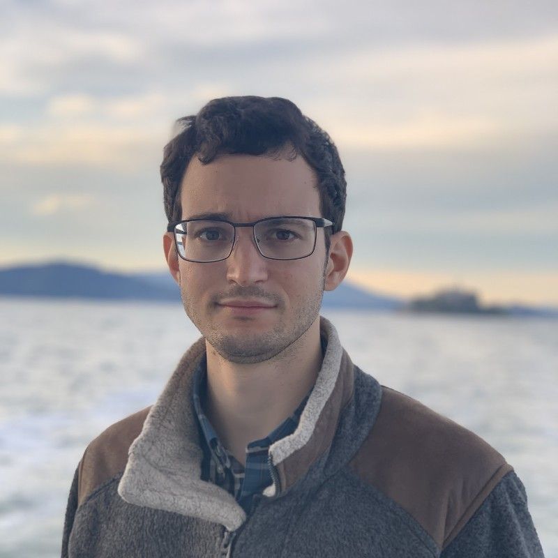 a man wearing glasses and a jacket is standing in front of a body of water .