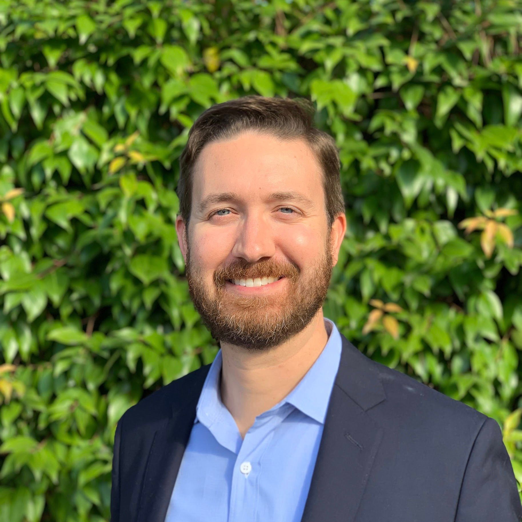 a man in a suit and blue shirt is smiling in front of a green bush .