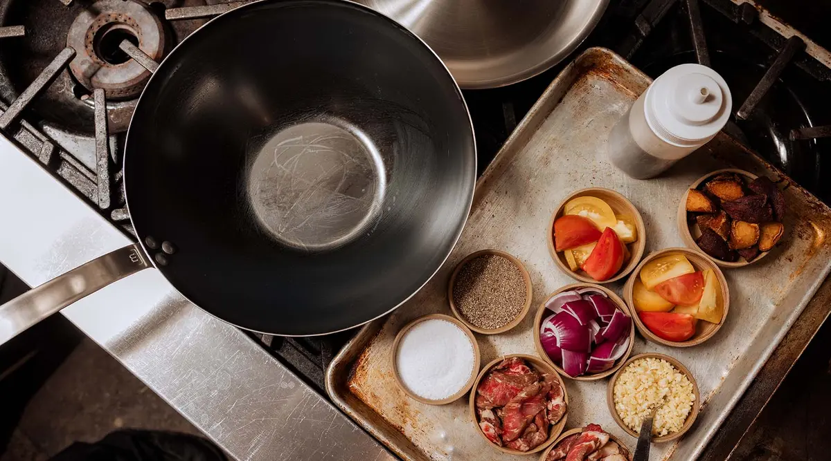 Image of Carbon Steel Wok on a stovetop next to stir fry ingredients that are in small bowls placed on a Sheet Pan.