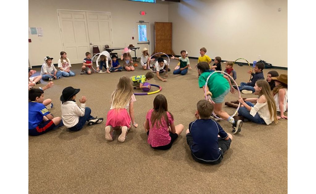 Campers sitting in a big circle enjoying a fun hula-hoop party game indoors.