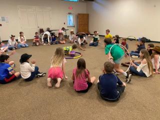 Campers sitting in a big circle enjoying a fun hula-hoop party game indoors.
