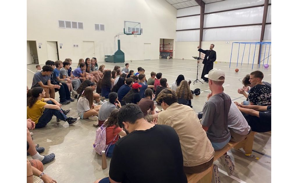 Father speaking to a big group of attentive campers gathered in the gym.