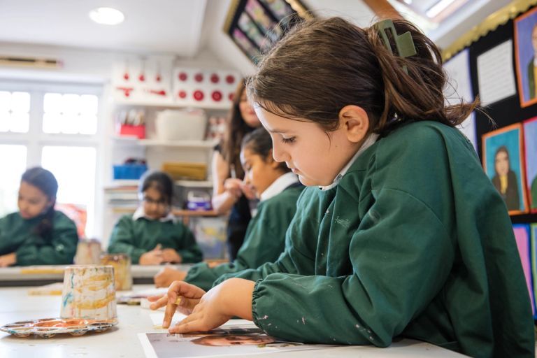 Girl in green smock focused on painting at a classroom table. Several other children, also in green smocks, are engaged in similar activities. A teacher is assisting them. The room is decorated with art supplies and colourful artwork on walls.