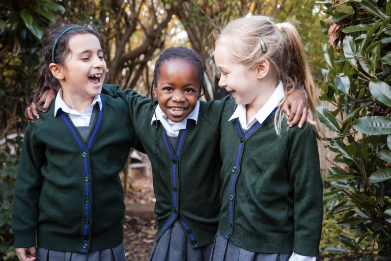 Three children in green Lyonsdown school uniforms smile and stand arm-in-arm outdoors. They are wearing white shirts, green cardigans with blue trim, and grey skirts. Surrounding them are trees and greenery, creating a friendly and joyful atmosphere.