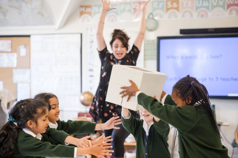 Children in Lyonsdown school uniforms eagerly catch a large cube passed by a teacher. The teacher, standing behind them, has arms raised in excitement. The classroom is bright, with educational posters and a screen in the background.