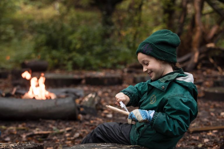A child in a green Lyonsdown jacket and beanie sits outdoors, smiling while carving a stick with a knife. A campfire burns in the background amidst a forest setting. The ground is covered with leaves and the atmosphere is cosy.