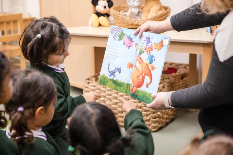 A teacher shows a colourful childrens book to a group of young students sitting on the floor. The book features a cartoon scene with animals and laundry on a line. The children are wearing green school uniforms.