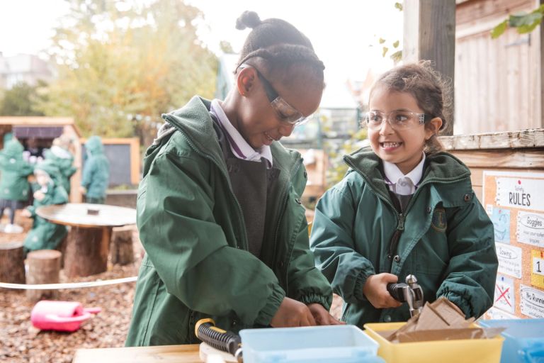 Two young children in green Lyonsdown uniforms and safety goggles work on a project outside. One is using tools, while the other smiles and watches. The background features a playground and greenery.
