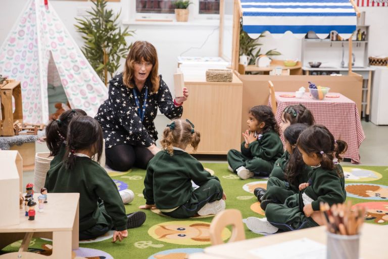 A teacher is sitting on the floor with a group of young children gathered in a circle on a colourful classroom rug. She is holding up a block and speaking to them. The classroom is equipped with play tents and various educational toys.