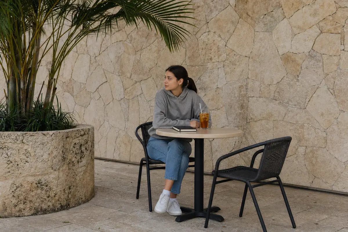 A young woman in a grey hoodie sits at a round café table on a shaded patio with an iced tea and a closed notebook, beside a stone planter.