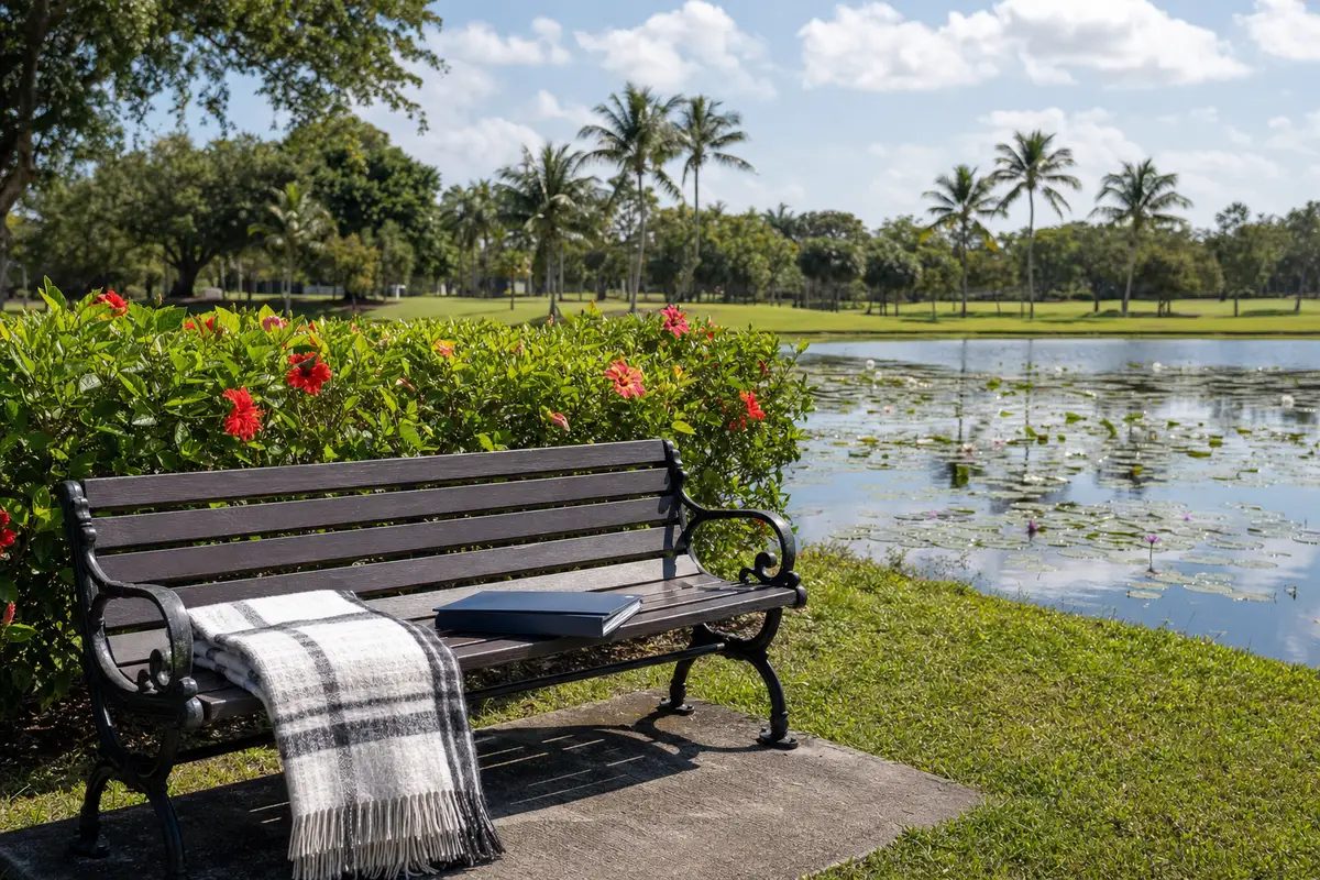 A wooden bench by a lake with a folded plaid blanket and a cushion, with flowering shrubs and palm trees behind it. No people are present.