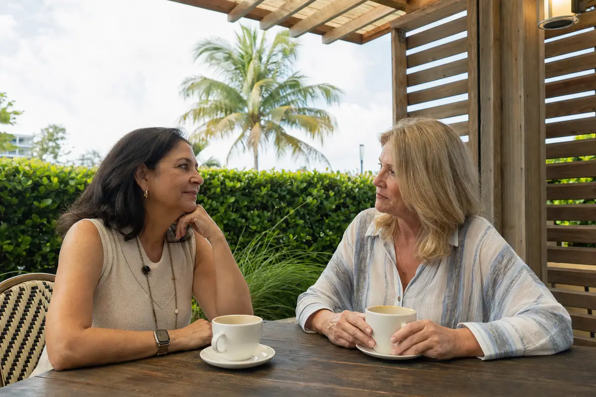 Two women talking over coffee at an outdoor table under a palm tree, with a leafy hedge behind them