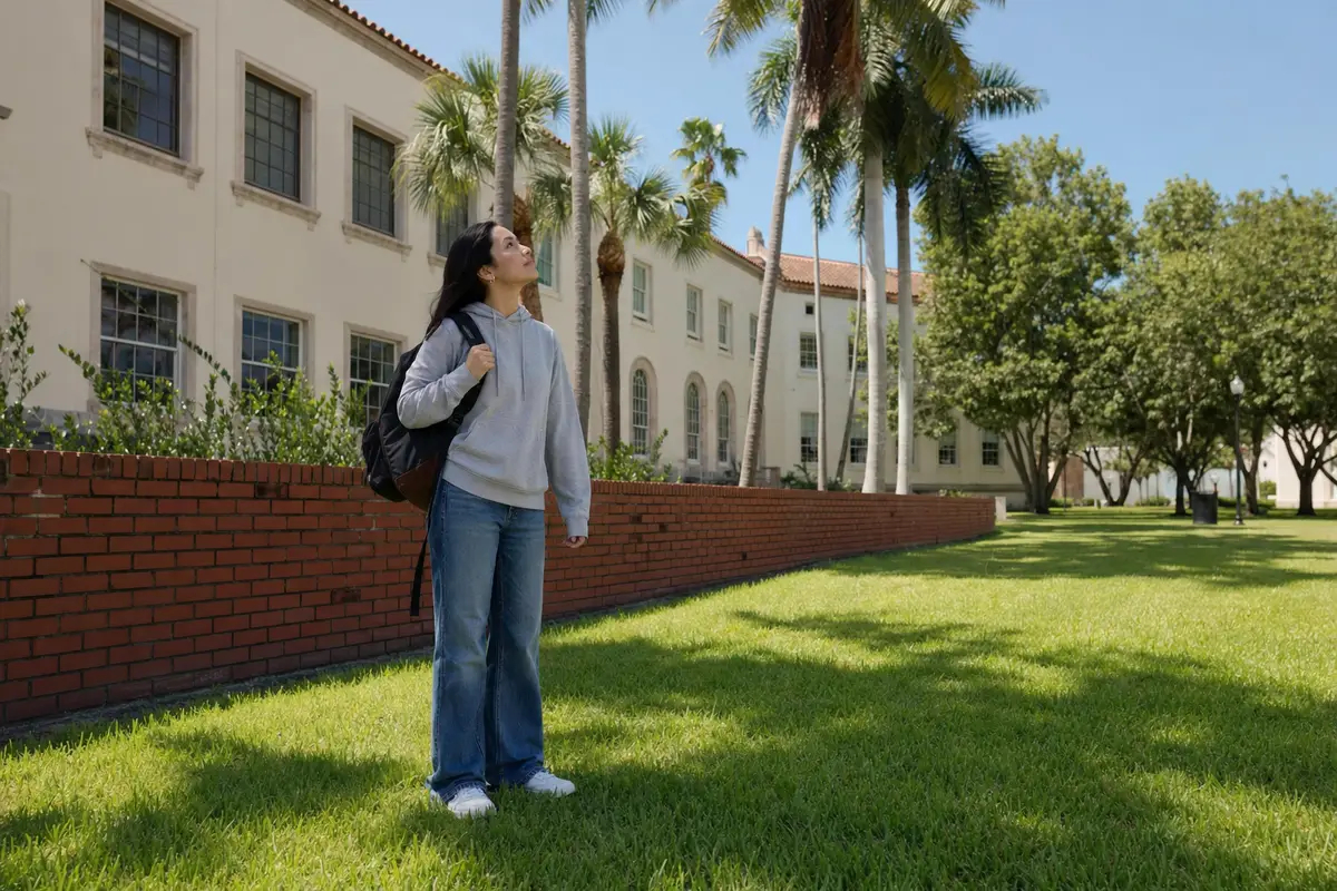 A young woman with a backpack stands on a campus lawn beside a brick wall and palm trees.
