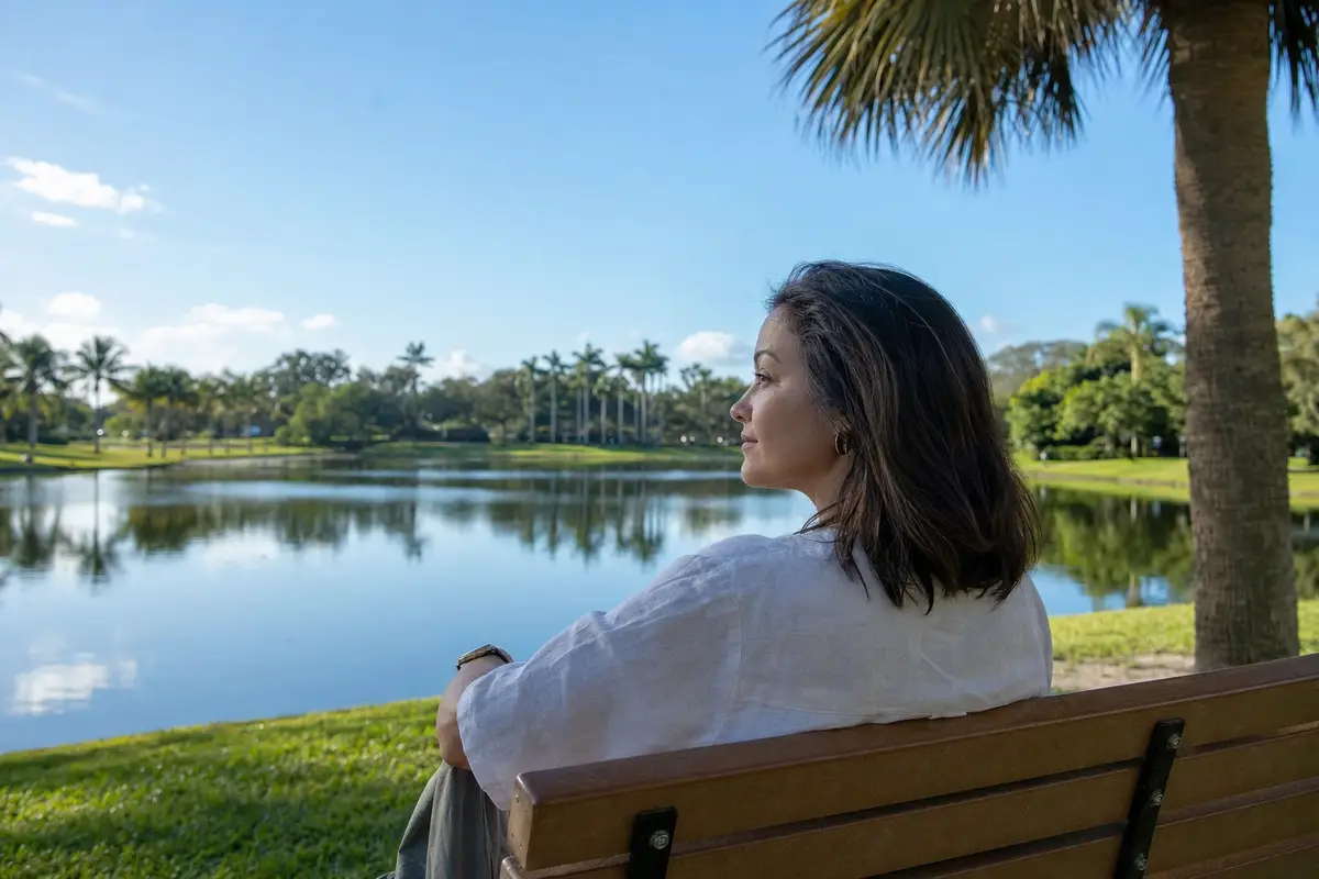 A woman sitting on a wooden bench by a calm lake, with palm trees on the far shore