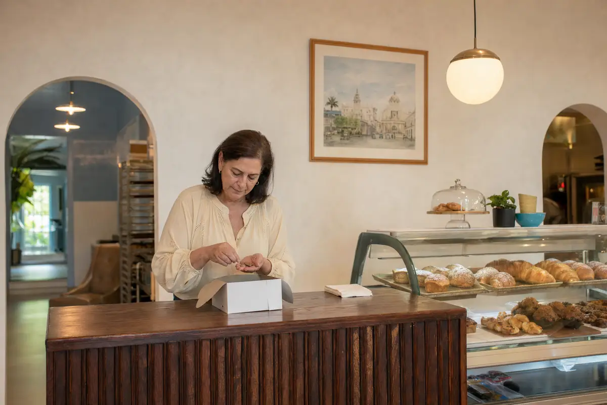 A woman in a cream blouse counts coins into her palm at a bakery counter beside a white paper box and a glass pastry case.