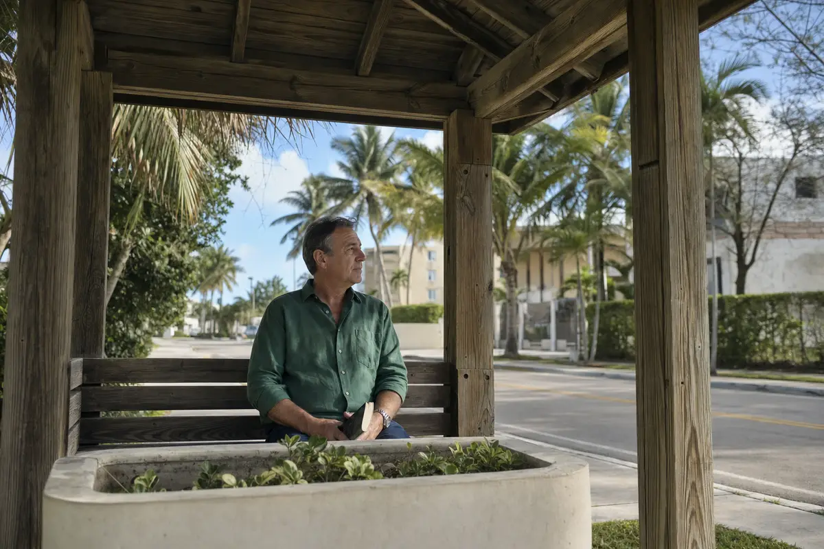 A man in a dark-green shirt sits on a wooden bench in a shaded bus-stop shelter, a closed paperback in his hands, looking out at a palm-lined street.