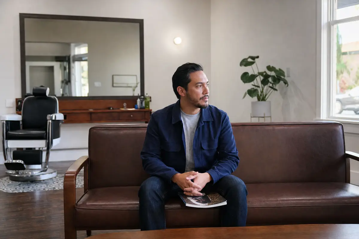 A man in a navy jacket sits on a brown leather bench beside a barber chair and a wall mirror, a magazine resting on his knee and his hands loosely clasped, looking calmly to the side.