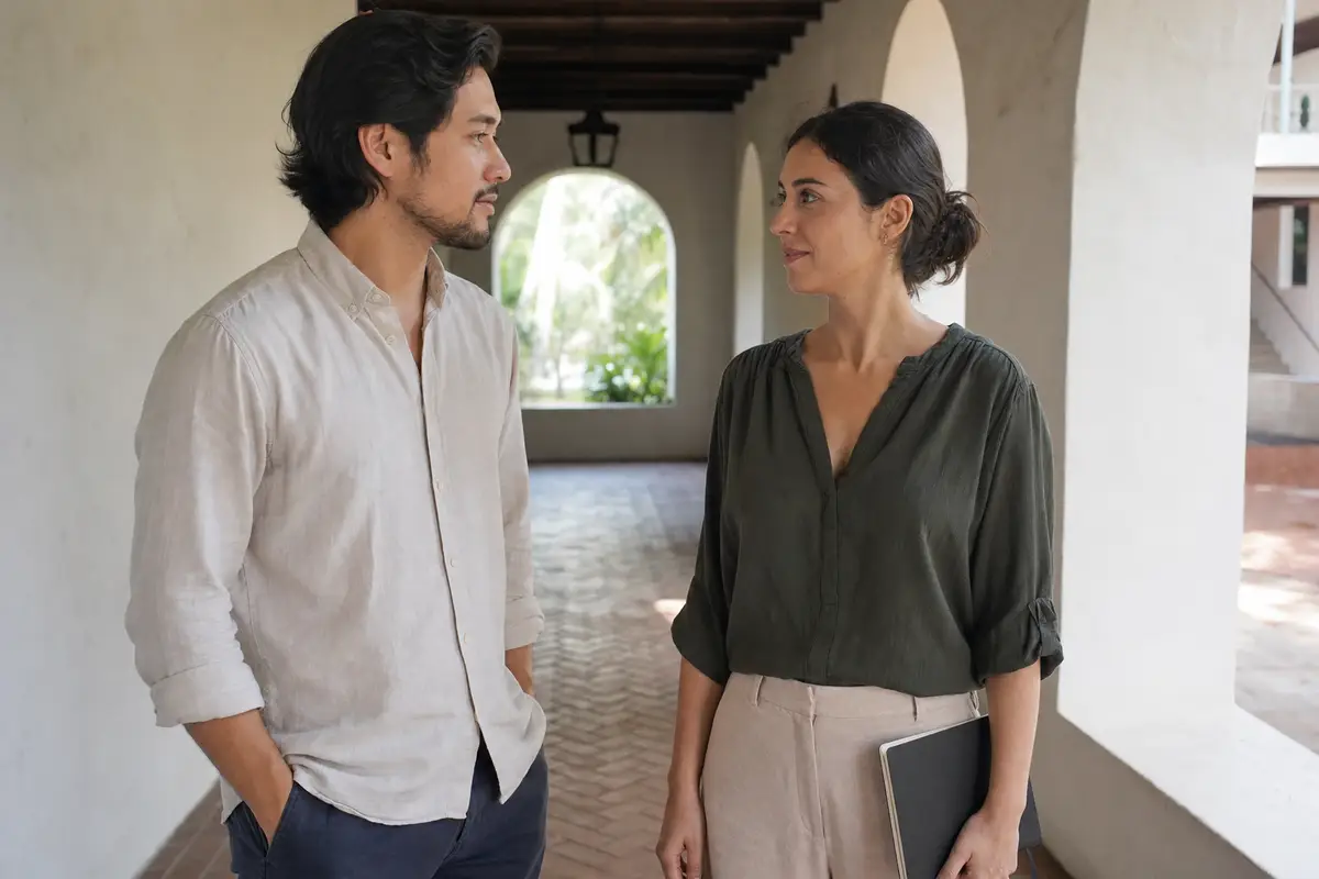 A man in a linen shirt and a woman in an olive top stand talking under an arched doorway in a courtyard.