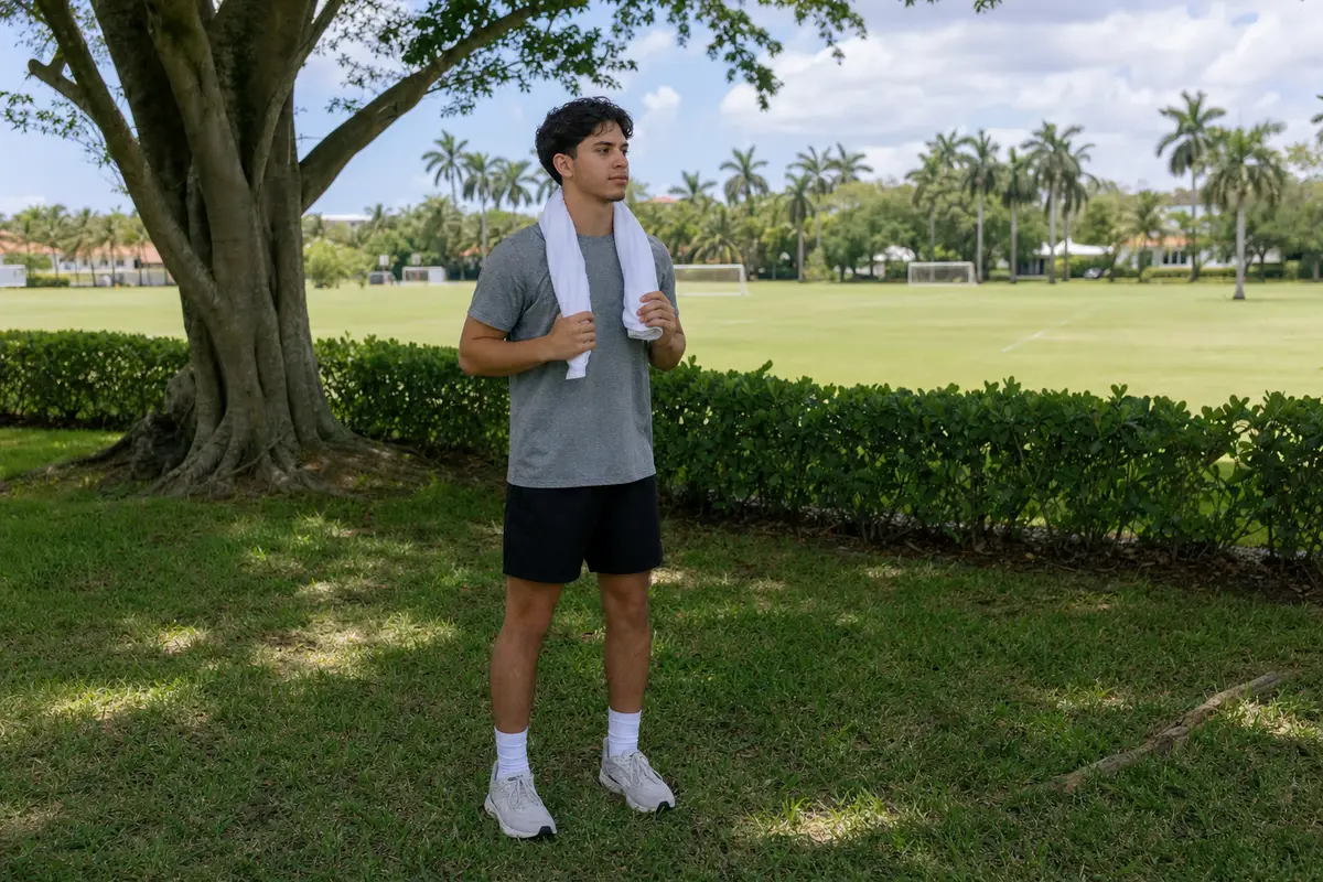 A young athlete in a grey shirt and black shorts stands on a grassy field beside a tree, holding a towel.