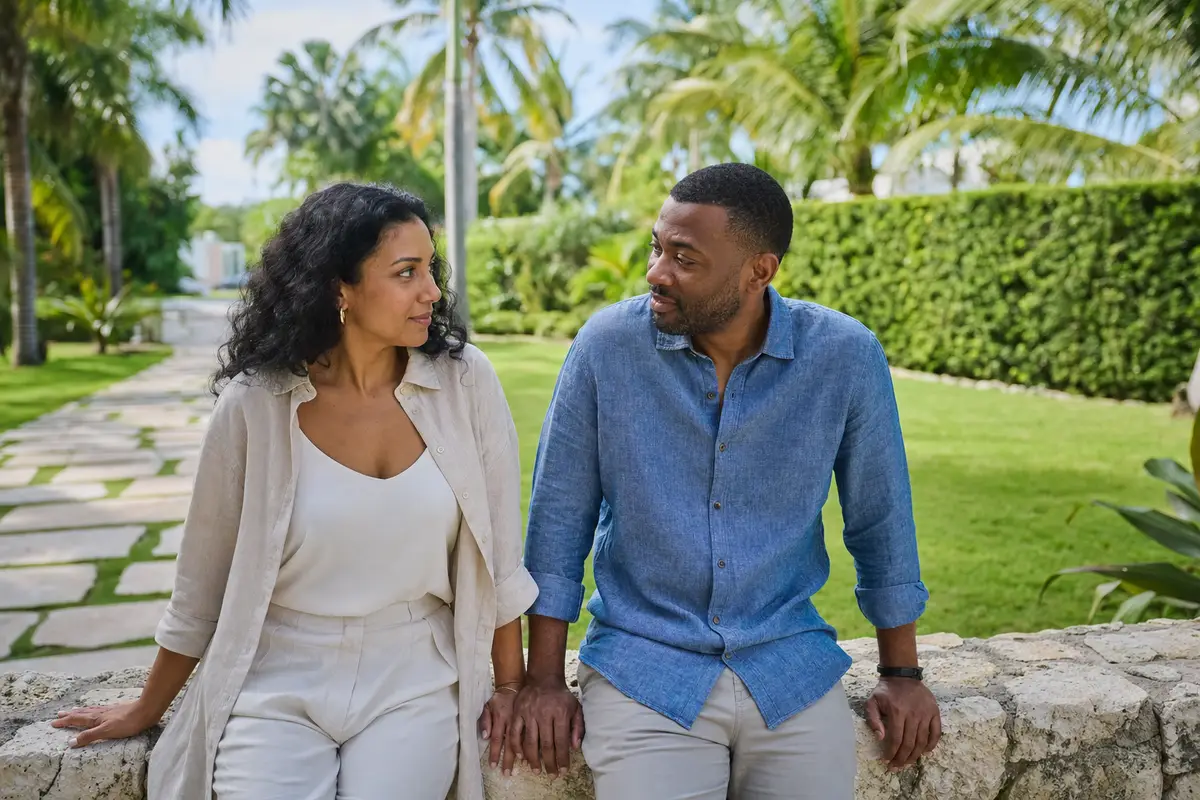 A couple sitting side by side on a stone garden wall under palm trees, talking calmly and looking at each other.