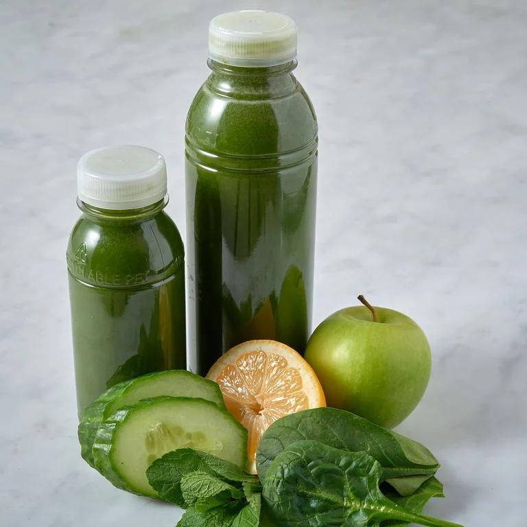 En liten och en större flaska med grön juice. Framför flaskorna ligger ett äpple, en näve spenat, en halv apelsin och två skivor gurka.  
